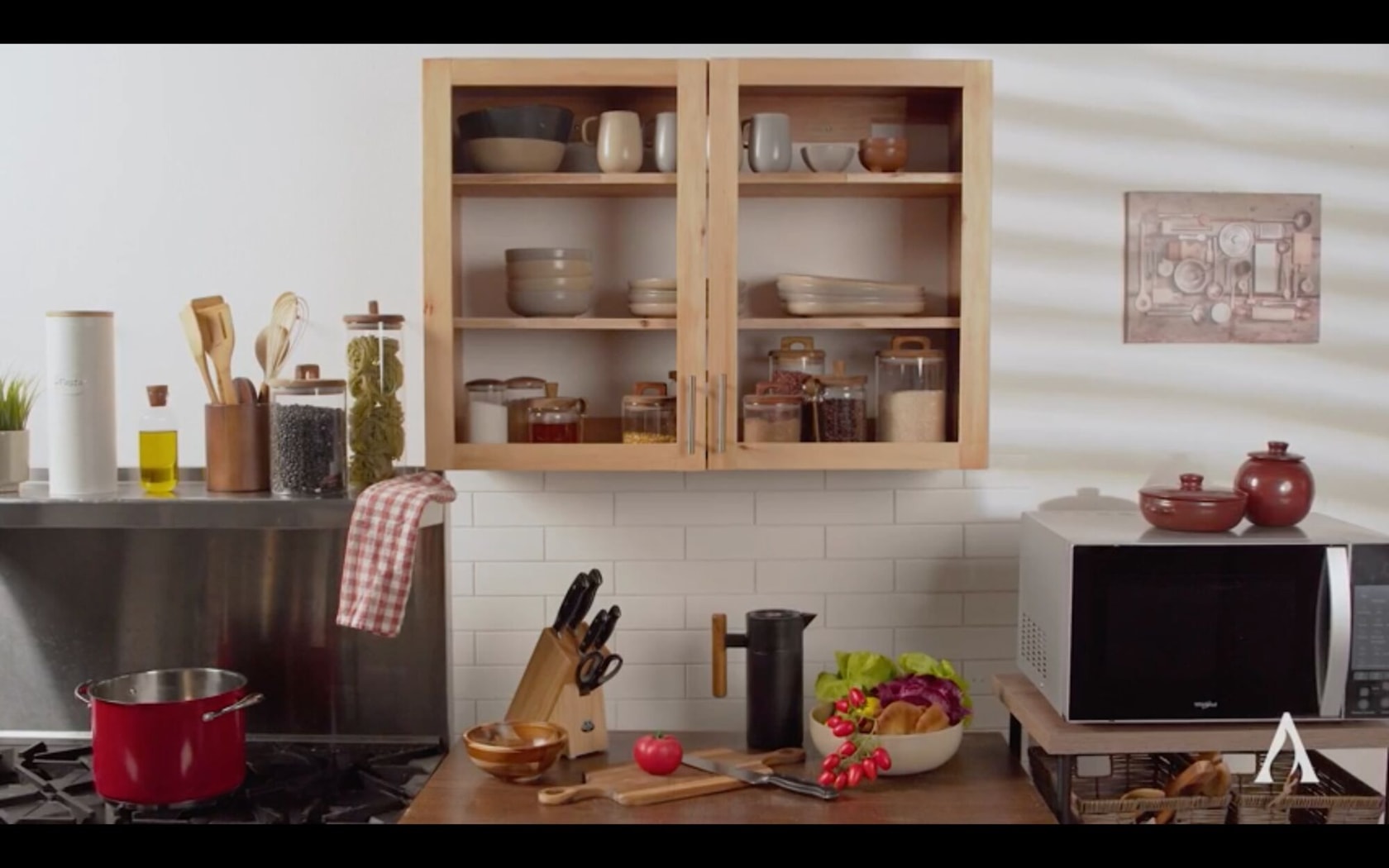 Una cocina moderna con gabinetes de madera, varios utensilios de cocina, frascos de ingredientes, una olla roja en la estufa, un bloque de cuchillos, una tabla de cortar con verduras, un microondas y recipientes de cerámica.