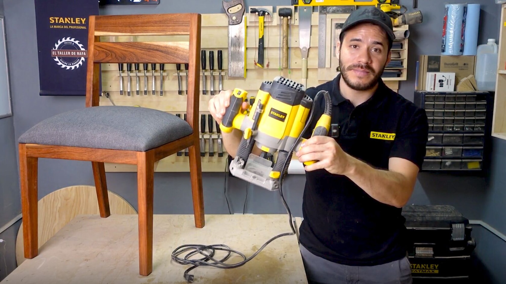 Una persona en un taller sostiene una herramienta eléctrica amarilla y gris de STANLEY, junto a una silla de madera sobre una mesa de trabajo. Hay varias herramientas manuales organizadas en la pared detrás.